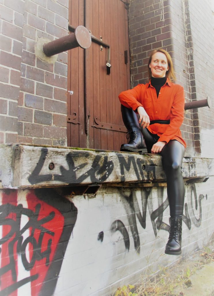 Juliane Mannteufel sitzt in einem orangefarbenen Oberteil vor einer Backsteinwand.